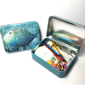 Puede incluir: Dos cajas met&aacute;licas rectangulares, una cerrada y otra abierta. La caja cerrada presenta una ilustraci&oacute;n de un pez azul con una peque&ntilde;a casa en su lomo. La caja abierta contiene varios suministros de manualidades, incluyendo ganchillos de metal, marcadores de puntos de pl&aacute;stico de colores y una herramienta circular blanca.
