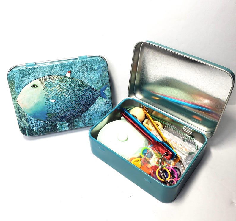 Puede incluir: Dos cajas met&aacute;licas rectangulares, una cerrada y otra abierta. La caja cerrada presenta una ilustraci&oacute;n de un pez azul con una peque&ntilde;a casa en su lomo. La caja abierta contiene varios suministros de manualidades, incluyendo ganchillos de metal, marcadores de puntos de pl&aacute;stico de colores y una herramienta circular blanca.