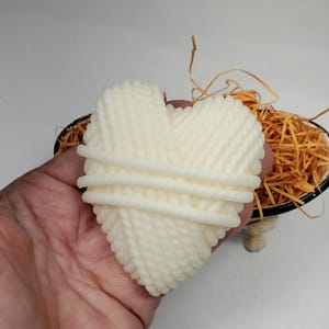 May include: A white heart-shaped yarn ball, resembling a skein of yarn, is held in a hand. The yarn ball is made of a soft, white material and has a textured surface.