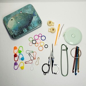 Puede incluir: Una caja de metal con una ilustraci&oacute;n de un pez azul, marcadores de puntos de pl&aacute;stico, dos cierres de cord&oacute;n beige, una aguja de pl&aacute;stico amarilla, una aguja de metal, una cinta m&eacute;trica redonda, tijeras de metal y ganchillos de crochet.