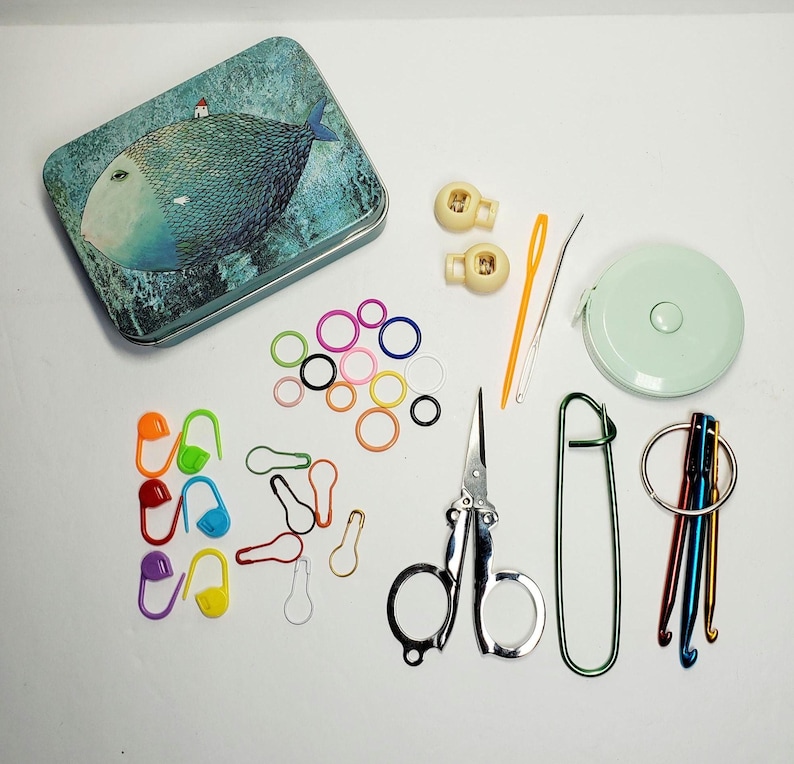 Puede incluir: Una caja de metal con una ilustraci&oacute;n de un pez azul, marcadores de puntos de pl&aacute;stico, dos cierres de cord&oacute;n beige, una aguja de pl&aacute;stico amarilla, una aguja de metal, una cinta m&eacute;trica redonda, tijeras de metal y ganchillos de crochet.
