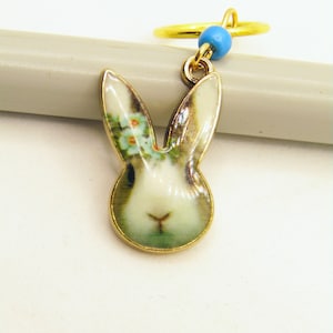 May include: A gold charm with a white rabbit face with blue flowers on its head. The charm is attached to a gold ring with a blue bead.