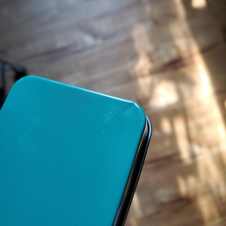 Puede incluir: Una caja met&aacute;lica rectangular de color verde azulado con esquinas redondeadas. La tapa presenta una superficie lisa y de color s&oacute;lido con un ligero brillo y una sutil marca de rasgu&ntilde;o cerca de la esquina superior derecha.