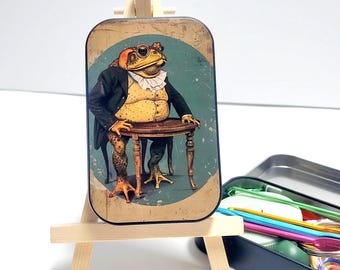 May include: A rectangular tin featuring a frog in a suit, seated at a table. The frog wears a black suit, a yellow vest, and a ruffled collar. The tin is displayed on a small wooden easel, with a black tin next to it.