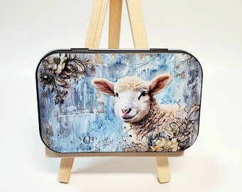 May include: A rectangular trinket box featuring a lamb illustration against a blue and white textured background with floral accents. The box is displayed on a small wooden easel, showcasing the artwork.