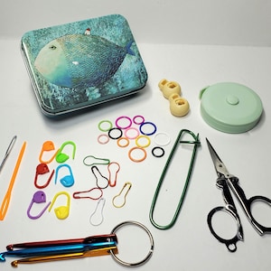 Puede incluir: Un kit de suministros para ganchillo y punto que incluye una lata rectangular con una ilustraci&oacute;n de un pez azul, una cinta m&eacute;trica verde menta, tijeras de metal, dos botones de pl&aacute;stico, un imperdible grande verde, marcadores de puntos coloridos y tres ganchillos de metal en una anilla.