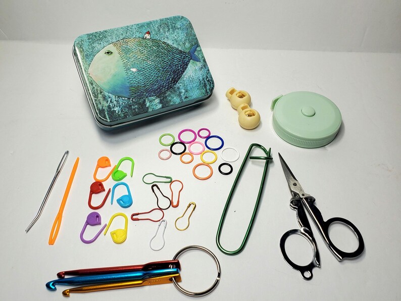 Puede incluir: Un kit de suministros para ganchillo y punto que incluye una lata rectangular con una ilustraci&oacute;n de un pez azul, una cinta m&eacute;trica verde menta, tijeras de metal, dos botones de pl&aacute;stico, un imperdible grande verde, marcadores de puntos coloridos y tres ganchillos de metal en una anilla.