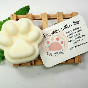 May include: A white paw-shaped beeswax lotion bar on a wooden stand with a label that reads "Beeswax Lotion Bar TOE BEANS Contains beeswax, coconut oil, shea butter, cocoa butter, lanolin, and essential oil KnittyKittyNotions.etsy.com"