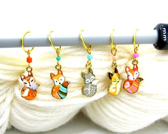 May include: Set of five gold-toned stitch markers with fox charms. Each fox charm is a different color and design, with colorful beads. The markers are on a knitting needle and white yarn.