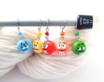 May include: A set of five colorful stitch markers, each featuring a round, smiling face design in blue, yellow, red, orange, and green. The markers are attached to a knitting needle, with a white yarn background.