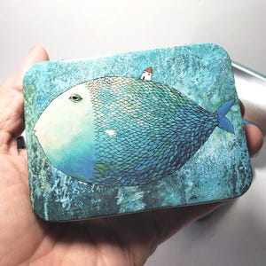 Puede incluir: Una lata rectangular de metal con la ilustraci&oacute;n de un pez grande, redondo, azul y verde con escamas detalladas. Una peque&ntilde;a casa blanca con techo rojo se asienta sobre el lomo del pez. El fondo es de color azul texturizado.