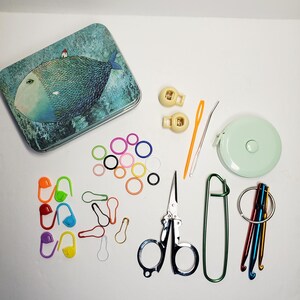 Puede incluir: Un conjunto de suministros de costura y ganchillo que incluye una lata rectangular con una ilustraci&oacute;n de un pez, dos cierres de cord&oacute;n beige, una aguja de pl&aacute;stico naranja, una aguja de metal, una cinta m&eacute;trica verde claro, marcadores de puntos coloridos, tijeras y ganchillos.