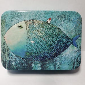 Puede incluir: Una caja de metal rectangular con una ilustraci&oacute;n caprichosa de un pez grande y redondo con cara p&aacute;lida, escamas azules y una peque&ntilde;a casa blanca con techo rojo sobre su espalda, sobre un fondo azul texturizado y moteado.