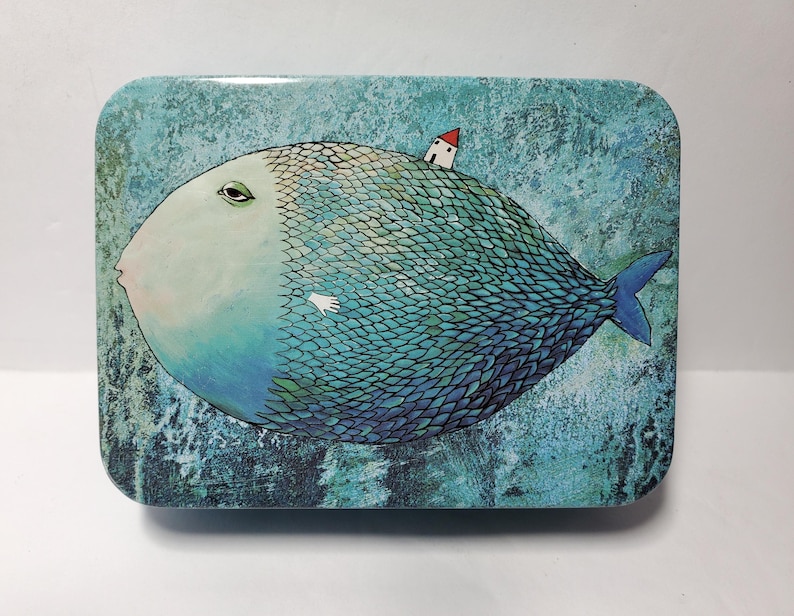 Puede incluir: Una caja de metal rectangular con una ilustraci&oacute;n caprichosa de un pez grande y redondo con cara p&aacute;lida, escamas azules y una peque&ntilde;a casa blanca con techo rojo sobre su espalda, sobre un fondo azul texturizado y moteado.