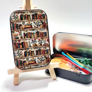 May include: A decorative rectangular tin with a bookshelf design on a small wooden easel. A black tin with craft supplies sits next to it, including crochet hooks and stitch markers. The bookshelf design features rows of books and floral accents.
