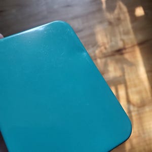 Puede incluir: Una caja de metal rectangular de color verde azulado s&oacute;lido con esquinas redondeadas. La superficie de la tapa es lisa y mate, presentando una ligera hendidura o abolladura cerca del borde superior derecho, dise&ntilde;o simple.
