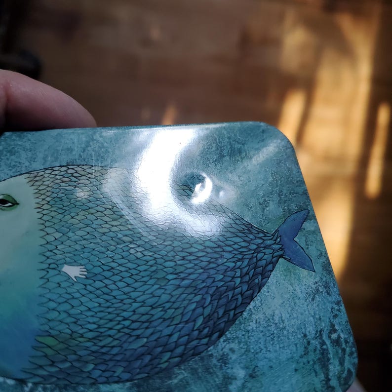 Puede incluir: Un posavasos rectangular con una ilustraci&oacute;n en azul verdoso y azul de un pez con un patr&oacute;n de escamas texturizado, un peque&ntilde;o detalle de mano blanca y una aleta caudal azul oscuro sobre un fondo azul verdoso.