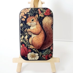 May include: A rectangular, decorative tin featuring a detailed illustration of a squirrel amidst a floral background. The squirrel is depicted in shades of brown and orange, surrounded by red, cream, and yellow flowers against a dark blue backdrop.