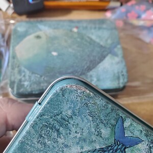 Puede incluir: Una caja de metal rectangular con una ilustraci&oacute;n texturizada en azul y verde azulado de un pez grande con una peque&ntilde;a casa blanca de techo rojo en su lomo. La tapa tiene un borde plateado y muestra signos de desgaste en el borde superior.