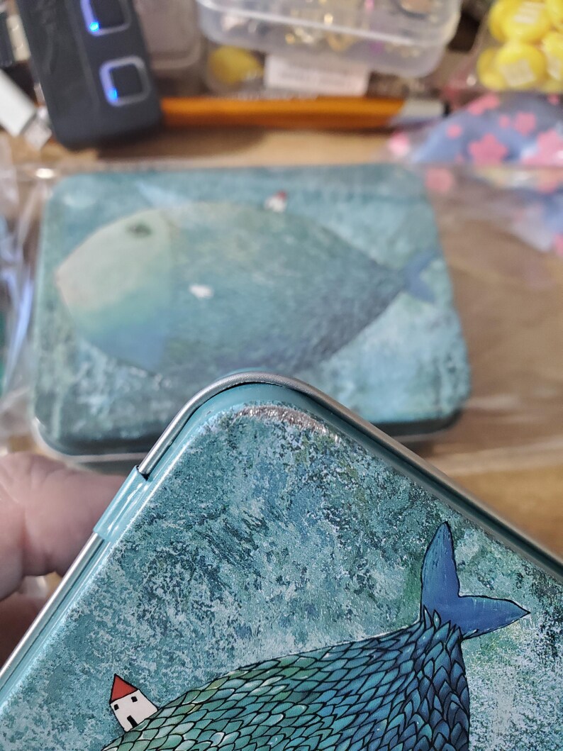 Puede incluir: Una caja de metal rectangular con una ilustraci&oacute;n texturizada en azul y verde azulado de un pez grande con una peque&ntilde;a casa blanca de techo rojo en su lomo. La tapa tiene un borde plateado y muestra signos de desgaste en el borde superior.