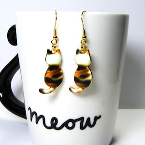 May include: A pair of cat-shaped earrings with gold-colored hooks. The earrings feature a white head, orange and black striped body, and a curved tail. The earrings are displayed on a white mug with the word "meow" in black script.