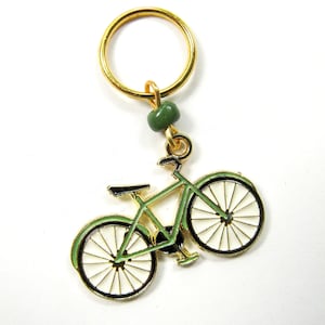 May include: A bicycle-shaped keychain with a gold-colored ring and accents. The bicycle is green with black and white details. A small green bead is attached to the top of the bicycle.