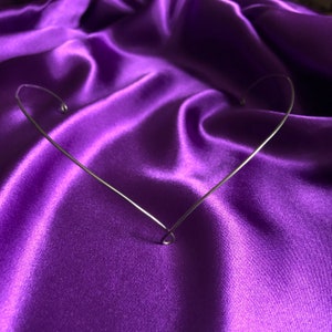 May include: A silver-colored wire headpiece shaped like a heart, resting on a vibrant purple satin fabric. The heart-shaped design is simple and elegant, with a delicate, curved wire construction. The background is a rich, textured purple.