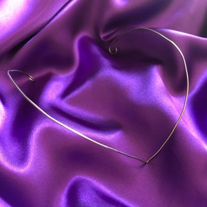 May include: A delicate silver-toned wire choker necklace in a heart shape, resting on a vibrant purple satin fabric. The necklace features a simple, elegant design with a curled end detail.