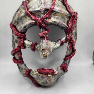 May include: A weathered mask with a skeletal appearance, featuring a textured surface in shades of gray and off-white. Bright red, vein-like structures wrap around the mask's contours, adding a striking contrast. The mask has a central nose-like projection and a wide, open mouth.