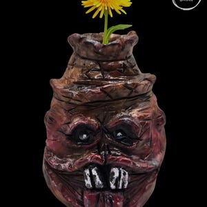 May include: A unique, handmade ceramic vase with a grotesque face design. The vase is painted in shades of brown and red, with a yellow flower in the opening. The vase has a textured surface and a dark background.