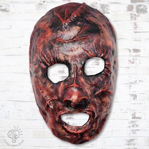 May include: A textured, full-face mask with a distressed appearance. The mask features a color palette of red, brown, and black, with open eye and mouth holes. The mask is likely for costume or theatrical use.