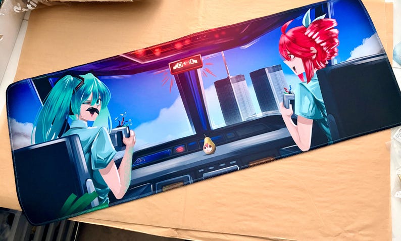 Op de afbeelding: Een rechthoekige muismat met een illustratie in anime-stijl. Het ontwerp bevat twee vrouwelijke personages met kleurrijk haar, tegen een achtergrond van een stadsgezicht en een blauwe lucht. De muismat is voornamelijk blauw en zwart.