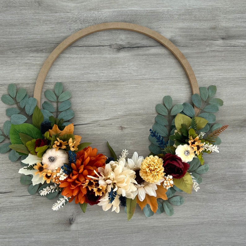 Puede incluir: Una corona de oto&ntilde;o con base de madera marr&oacute;n, decorada con flores artificiales en tonos naranja, rojo, blanco y amarillo. La corona tambi&eacute;n incluye hojas verdes y peque&ntilde;as calabazas blancas.