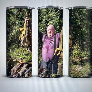 May include: A stainless steel tumbler with a colorful image of a man holding a large lizard. The image is set against a backdrop of green foliage.