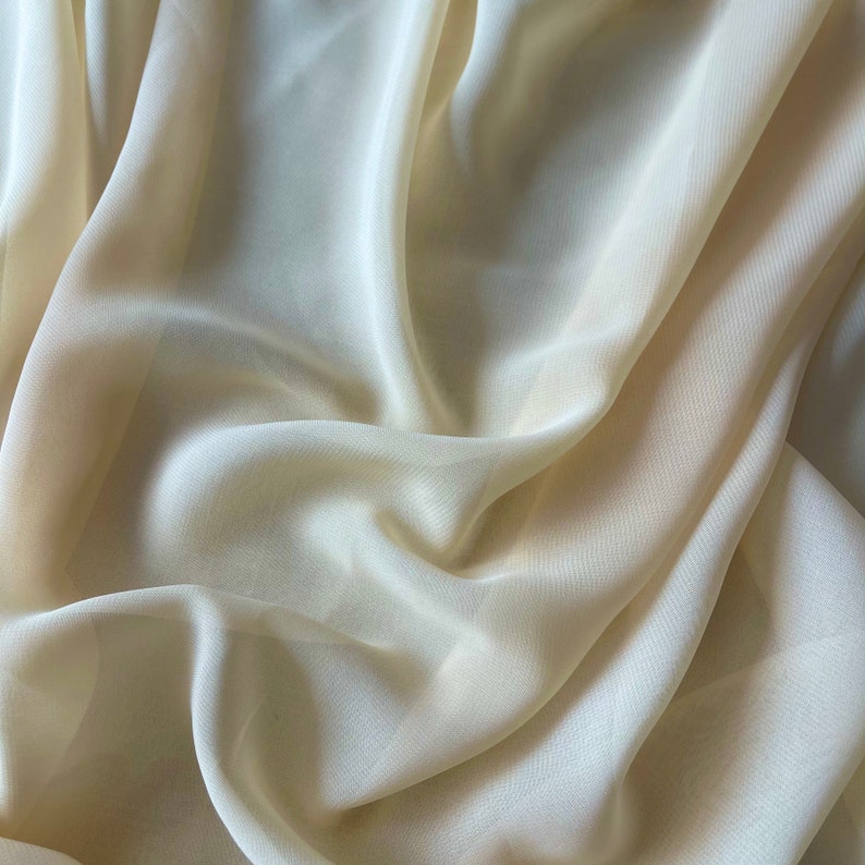 K&ouml;nnte beinhalten: Nahaufnahme eines weichen, hellgelben Stoffes mit glatter, seidenartiger Textur. Der Stoff ist drapiert und gefaltet und erzeugt einen flie&szlig;enden, eleganten Look.