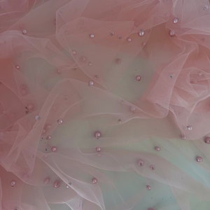 May include: Pink tulle fabric with scattered pearl-like embellishments. The fabric is draped and folded, creating a soft and delicate texture.