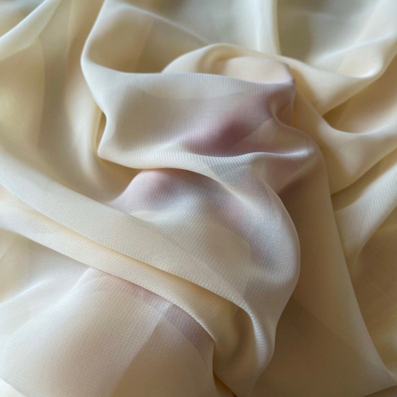 K&ouml;nnte beinhalten: Nahaufnahme eines weichen, cremefarbenen Stoffes mit einer dezenten Textur. Der Stoff ist drapiert und gefaltet, wodurch ein glatter, flie&szlig;ender Look entsteht.