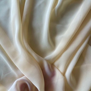 K&ouml;nnte beinhalten: Nahaufnahme eines weichen, cremefarbenen Stoffes mit einer glatten, seidenartigen Textur. Der Stoff ist drapiert und gefaltet, was einen luxuri&ouml;sen und eleganten Look erzeugt.