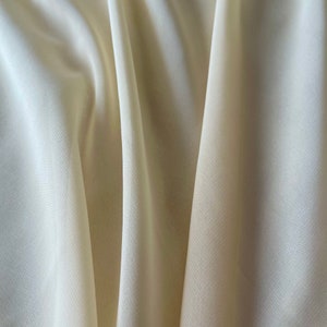 K&ouml;nnte beinhalten: Nahaufnahme eines glatten, elfenbeinfarbenen Stoffes mit einem dezenten Glanz. Der Stoff ist in weichen Falten drapiert und erzeugt einen luxuri&ouml;sen und eleganten Look.