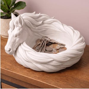 May include: A white horse head-shaped decorative bowl, ideal for holding keys and coins. The bowl is made of a solid material and features a detailed mane and a braided design around the rim. The bowl is sitting on a wooden surface.
