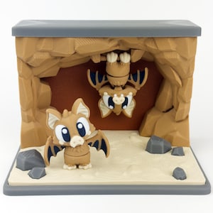 May include: A diorama featuring two cartoon bats in a cave setting. One bat stands upright, while another hangs upside down from the cave ceiling. The cave is brown with gray accents, and the bats have large eyes and outstretched wings.