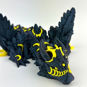 May include: A 3D-printed dragon figurine in dark blue and yellow. The dragon features intricate feather-like details and yellow accents around the head and body. The figurine is posed on a white surface.