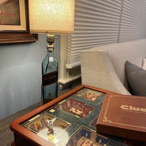 May include: A wooden game board with a glass top, featuring a miniature replica of a mansion. The board is labeled "Clue Luxury Edition". A lamp made from a repurposed bottle of "Johnnie Walker Blue Label" whiskey sits on a table next to the board.