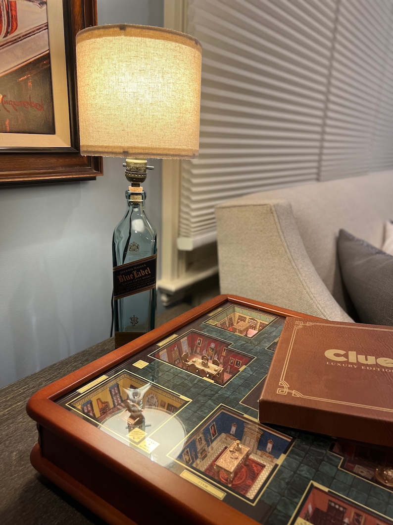 May include: A wooden game board with a glass top, featuring a miniature replica of a mansion. The board is labeled "Clue Luxury Edition". A lamp made from a repurposed bottle of "Johnnie Walker Blue Label" whiskey sits on a table next to the board.