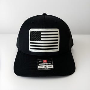 May include: Black trucker hat with a white and black American flag patch. The hat has a mesh back and a curved brim. The patch is rectangular and features the stars and stripes of the US flag. The hat has a Richardson 112 tag.