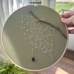 May include: A round embroidery hoop featuring a spider web design. The web is crafted with clear beads and attached to a small twig. A black spider hangs from the web. The fabric is a muted green color, and the hoop is held by a hand.