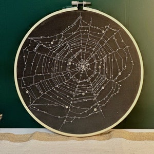 May include: A black embroidery hoop with a white spider web design. The web is decorated with small, white beads.