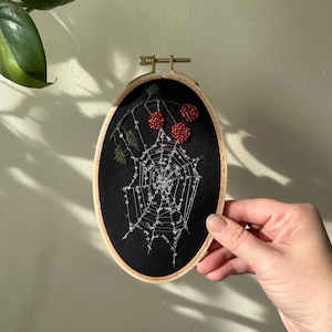 May include: Oval embroidery hoop featuring a black fabric with a detailed spiderweb design. Red beaded berries and green leaves are embroidered at the top. The wooden hoop is held by a hand.