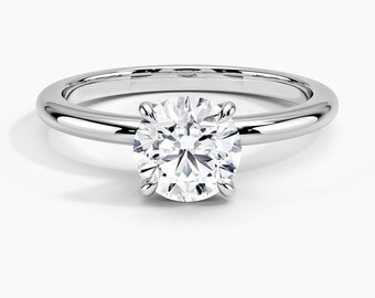 May include: A silver-toned metal ring featuring a single round-cut clear gemstone held in a four-prong setting. The band is smooth, polished, and circular, showcasing a classic design with a brilliant, faceted center stone that reflects light.