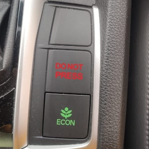May include: A close-up of a car's dashboard with a black button that says "DO NOT PRESS" in red text and a green button with a leaf icon and the word "ECON" in white text.